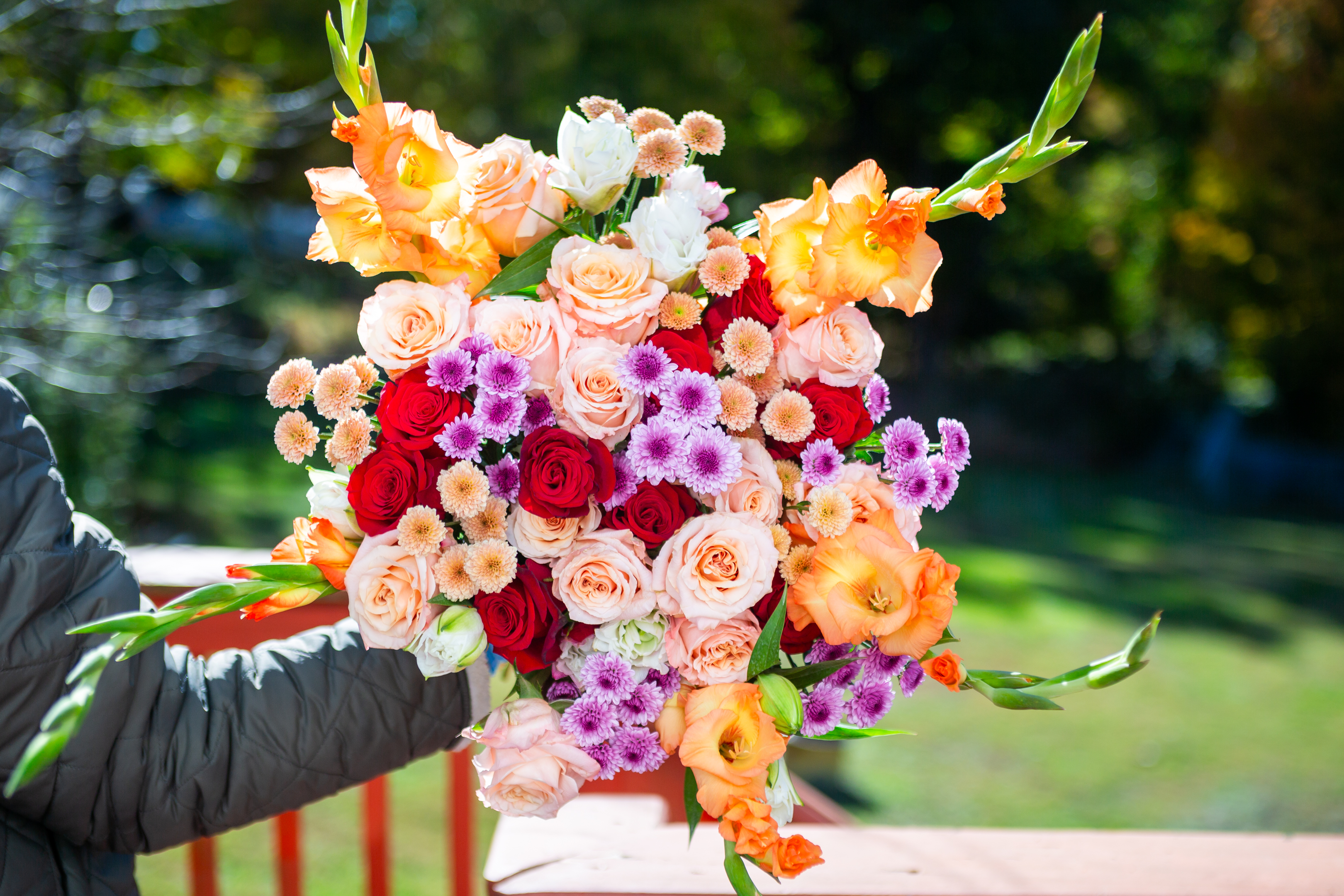 Jellybean — a colorful hand-tied bouquet of gladiolus, blush and red roses, and lavender mums held outdoors, Livingston NJ florist